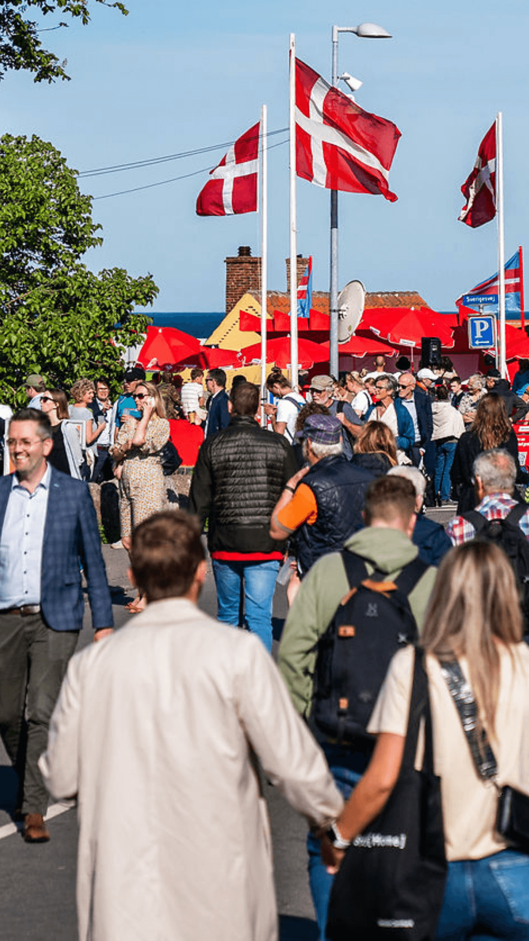 a crowd of people are walking down a street with flags flying in the background at Folkemødet on Bornholm