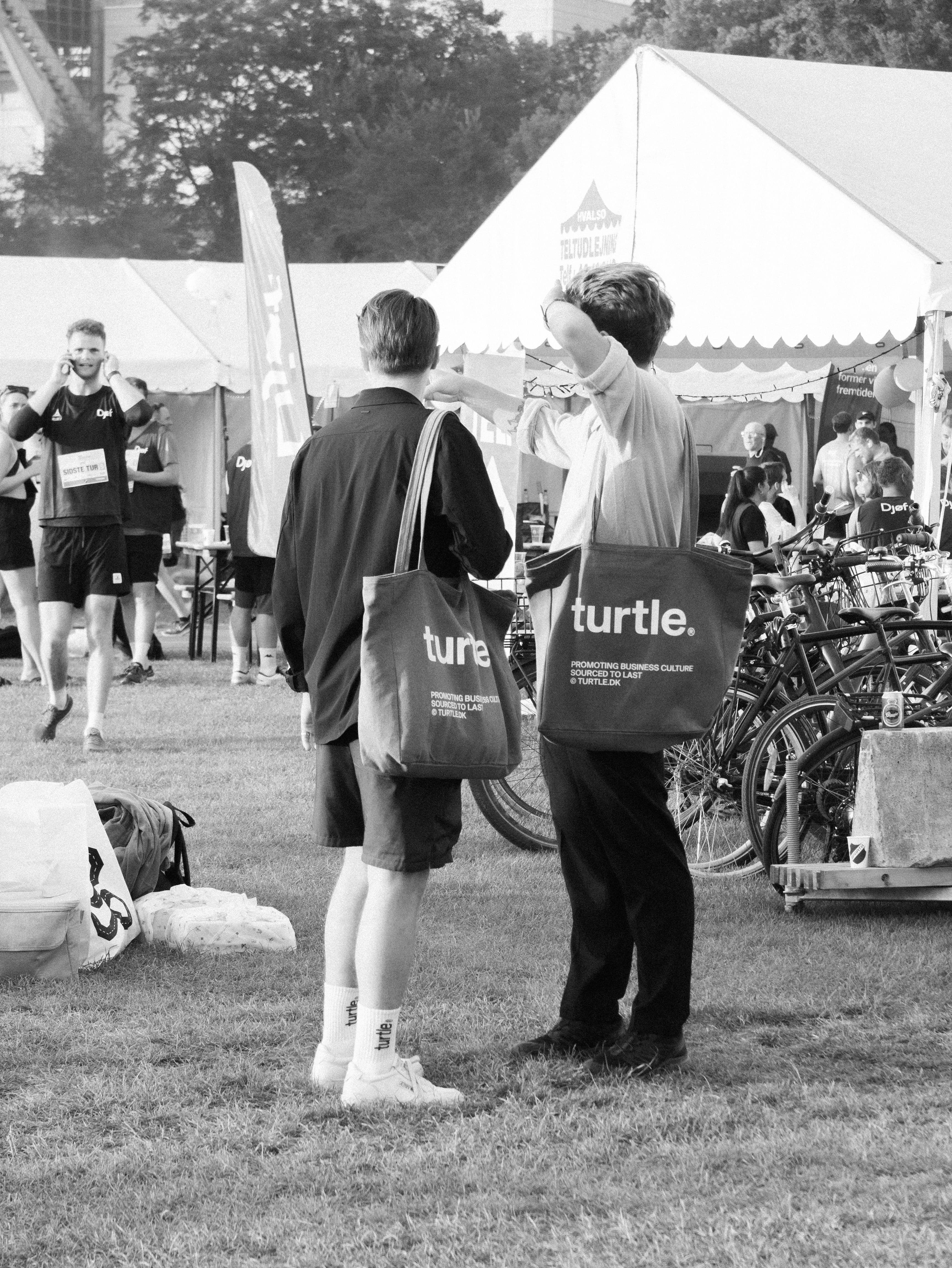 Black and white photo of two men with 'turtle.' tote bags at a grassy outdoor event with tents and people.