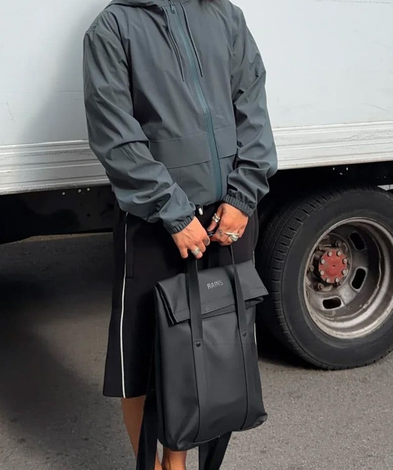 A person in a grey hooded jacket and black pants holds a black Rains roll-top backpack.